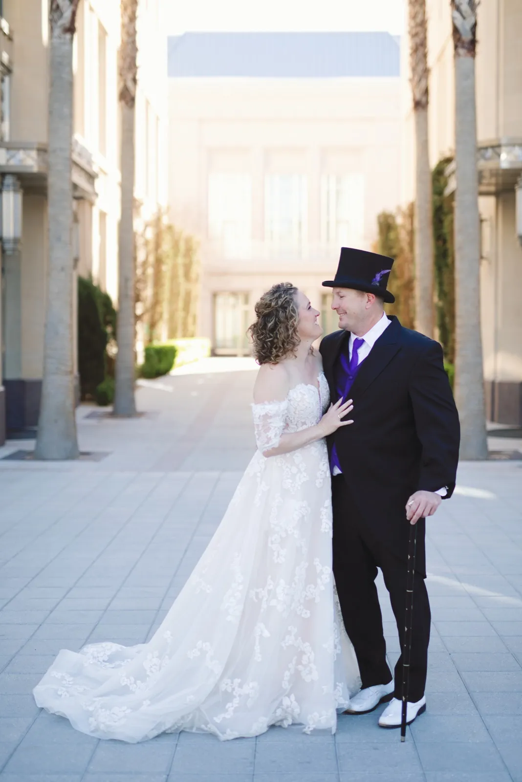 Couple standing face to face in a quiet palm lined courtyard, her long lace train spread across the pavement
