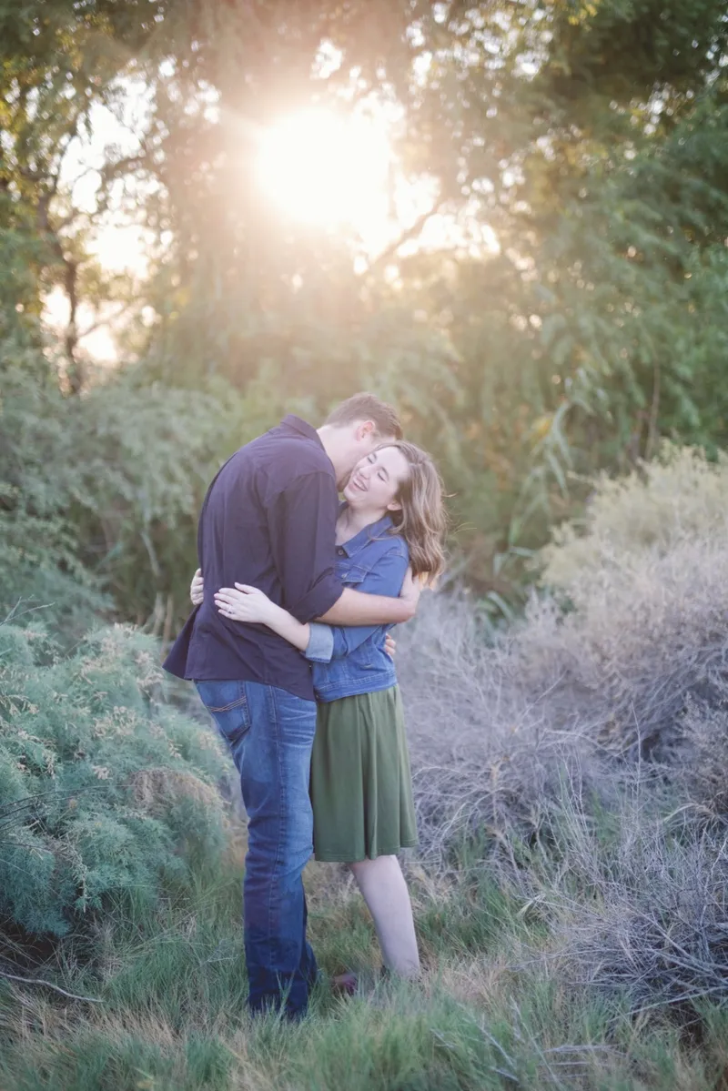 Couple holding each other under a tree with the sun flaring through the branches and pale desert brush all around them