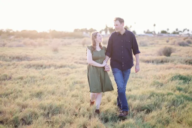 Couple holding hands and smiling at each other as they walk through a wide field of tall grass with the sun low behind them