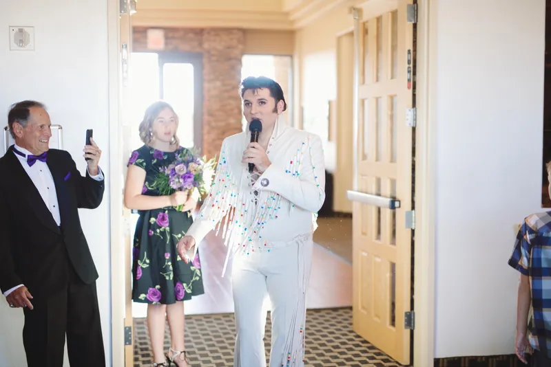 An Elvis performer in a white jumpsuit singing into a microphone in the clubhouse doorway while two guests wait beside him
