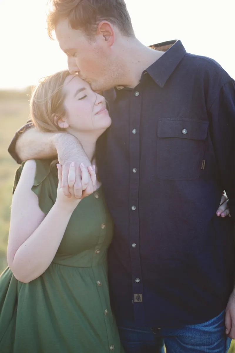 Man kissing the top of his fiancee's head while she holds onto his arm across her shoulder