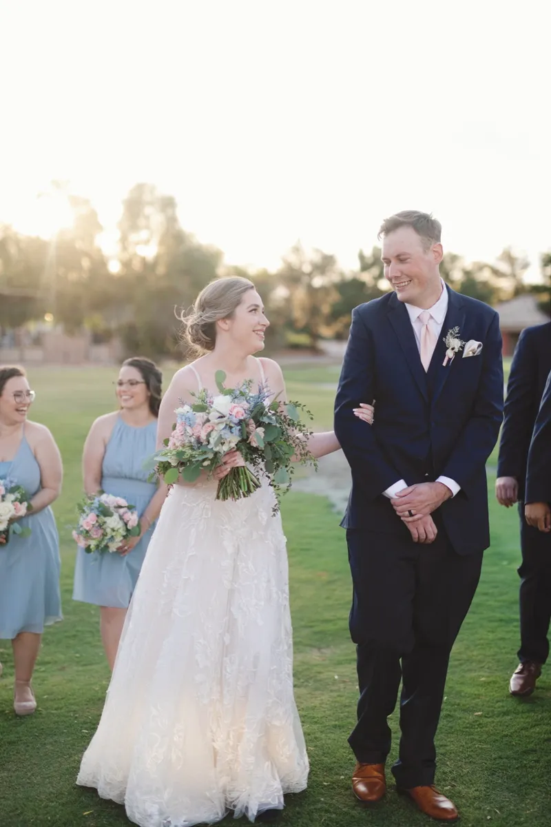 Couple walking across the lawn with bridesmaids in dusty blue holding pastel bouquets alongside them