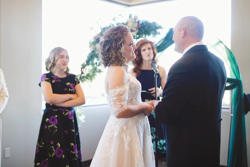 Couple holding hands through their vows in front of a greenery arch with a bridesmaid beside them