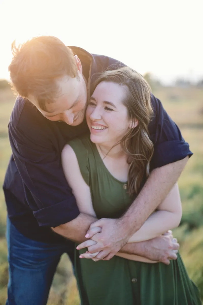 Man leaning over his fiancee's shoulder with his arms wrapped around her, both of them laughing in warm backlight