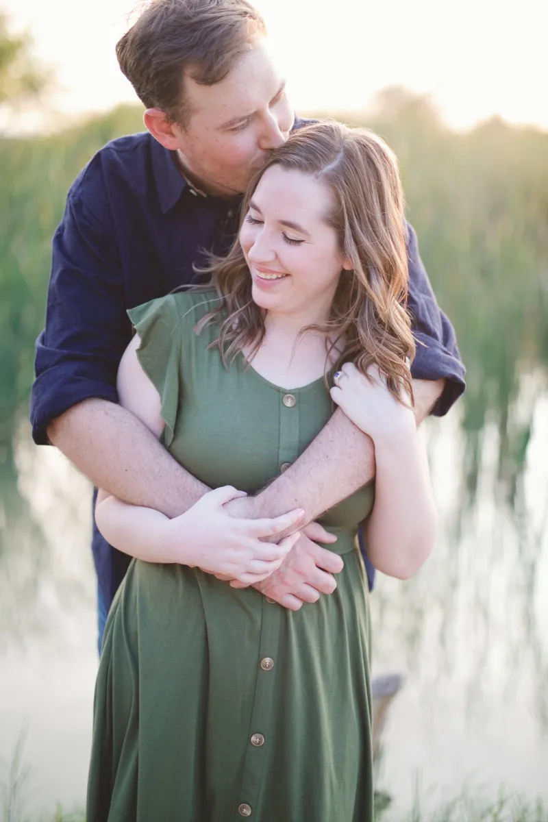 Man with his arms around his fiancee kissing the side of her head at the edge of still water with tall reeds behind them