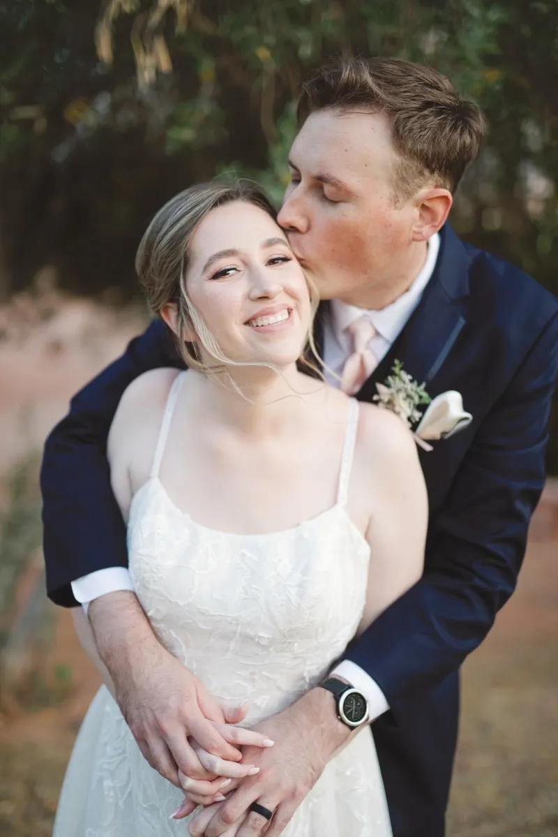 Groom kissing the bride's cheek outdoors while she smiles at the camera