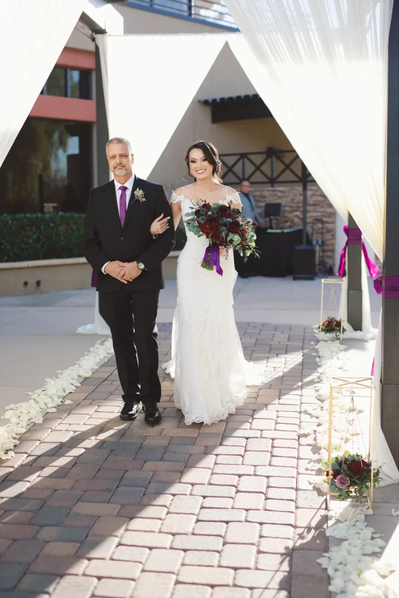 Bride walking in on an older man's arm along the paver aisle under the patio drapes, her bouquet in deep purple and burgundy