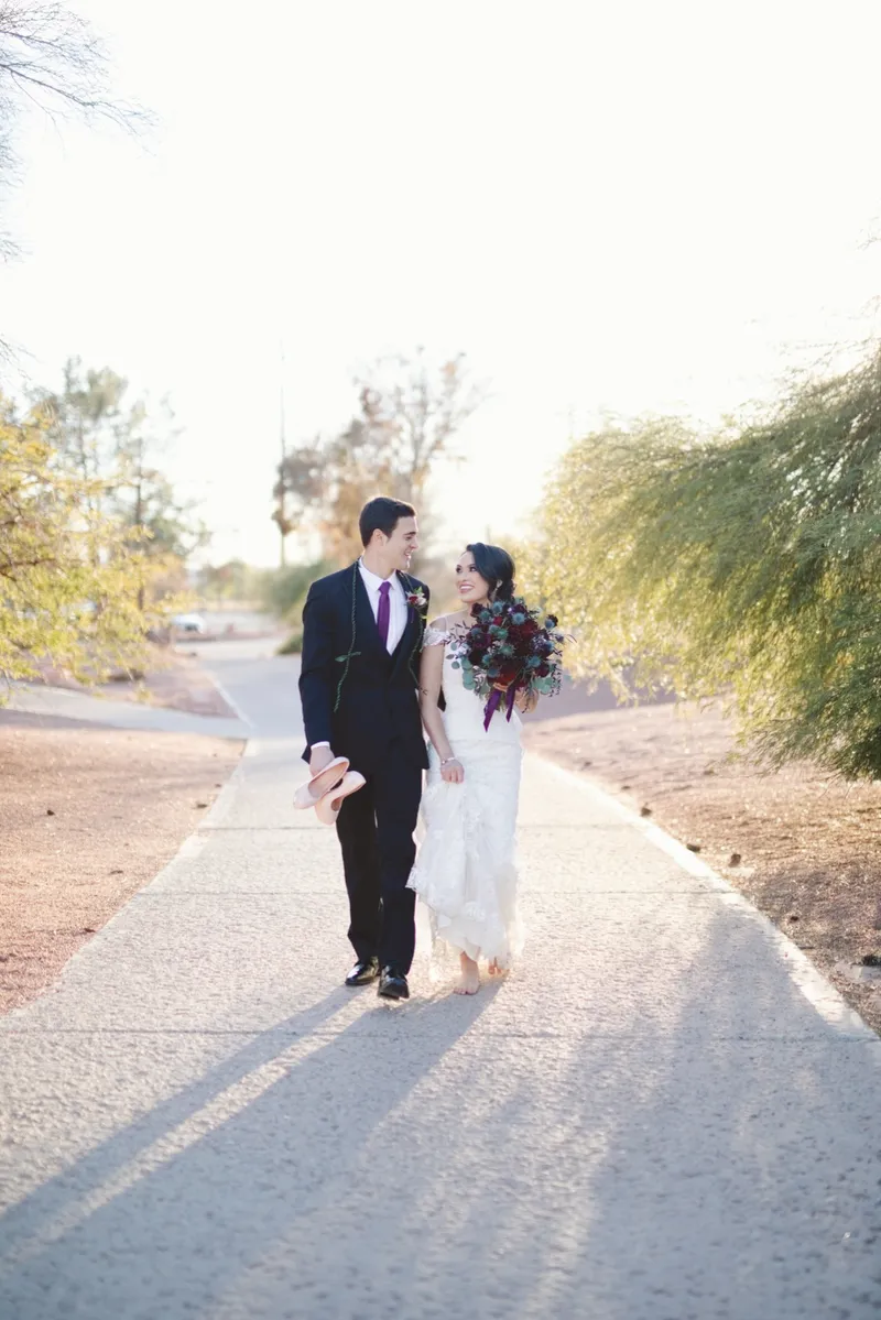 Couple walking a shaded path together, the groom carrying the bride's heels in one hand