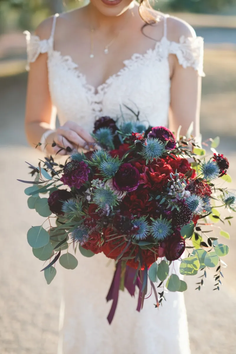 Close crop of a bouquet of burgundy roses, blue thistle and eucalyptus held against a white gown