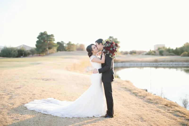 Groom kissing the bride beside the pond on the course in low golden light with her burgundy bouquet raised