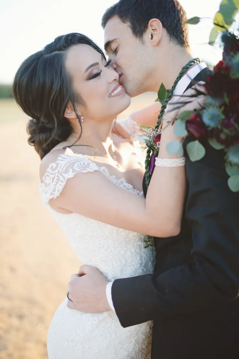 Close portrait of the groom kissing the bride's cheek with her burgundy bouquet raised beside them