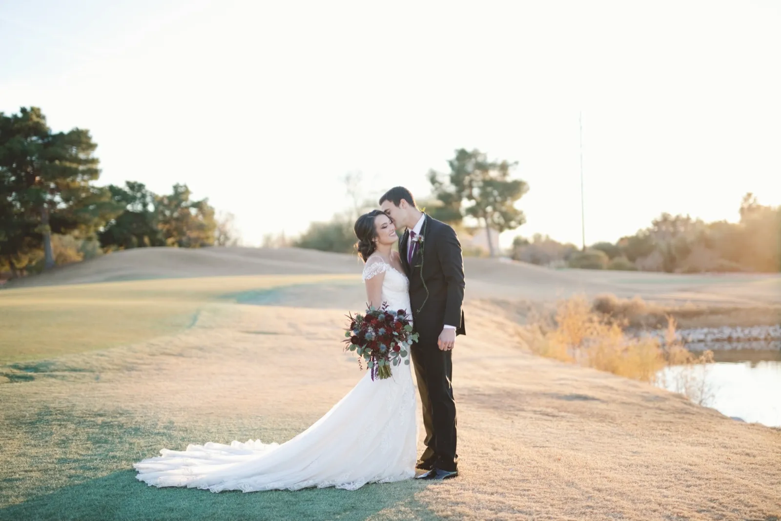 Couple stand close on the fairway at golden hour with a pond beside them and the sun low