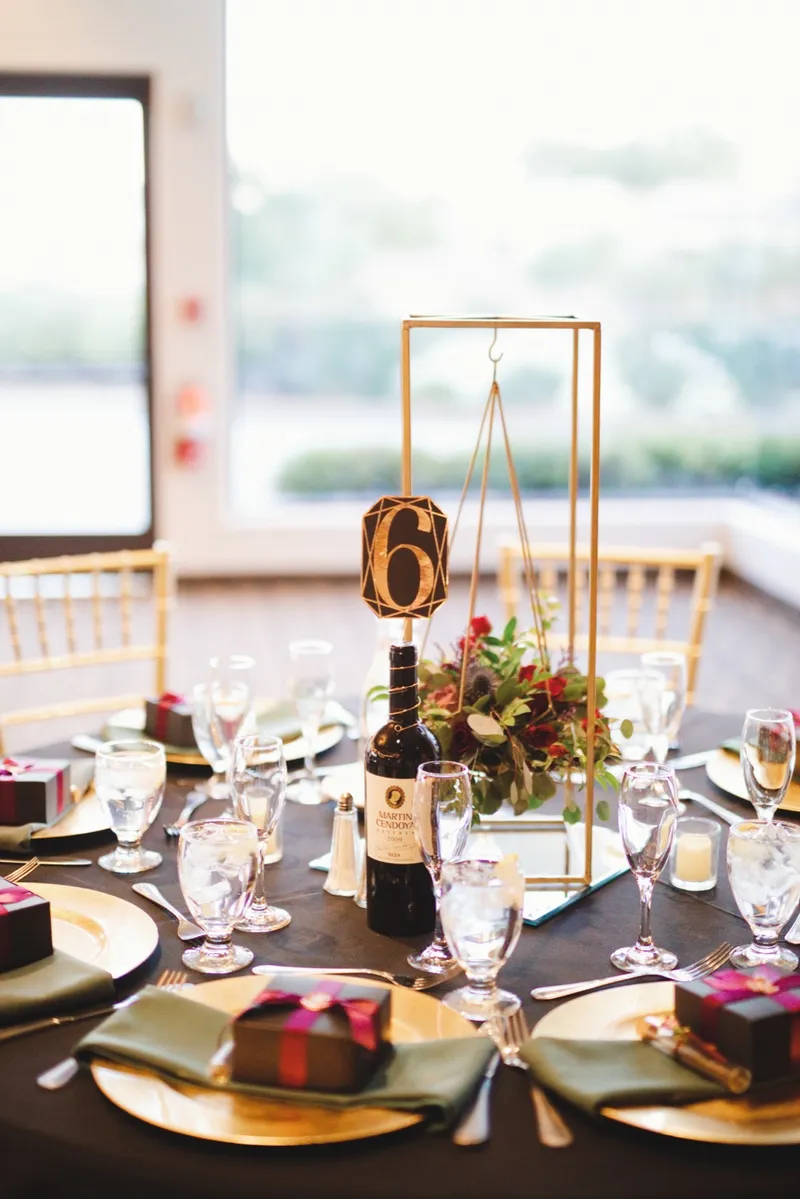 Table six marker in a gold frame with a wine bottle and burgundy florals beside the place settings