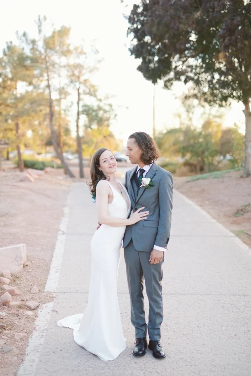 Couple standing together on the path, the groom in a grey suit with his arm around her waist