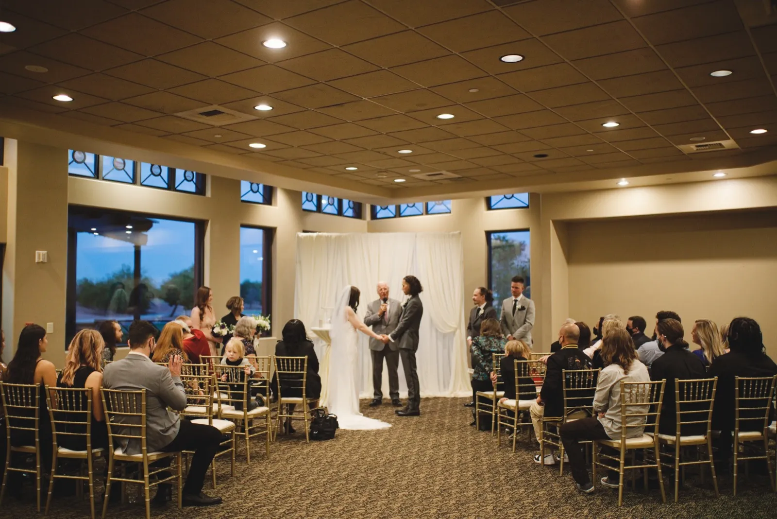Indoor ceremony at a white draped backdrop, guests on gold chairs, windows down two walls