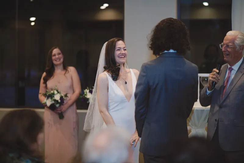 Bride laughing at her partner during the indoor ceremony while the officiant and a bridesmaid stand beside them