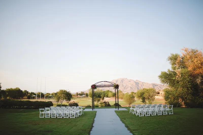 Empty ceremony lawn with two blocks of white folding chairs facing a black iron rotunda topped with florals, the golf course and a long mountain ridge behind it