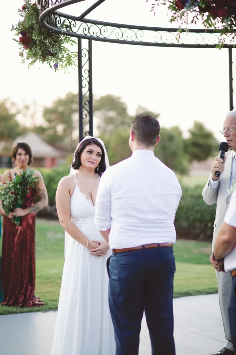 Couple standing under the iron rotunda with the officiant holding a microphone beside them and a bridesmaid watching from the grass