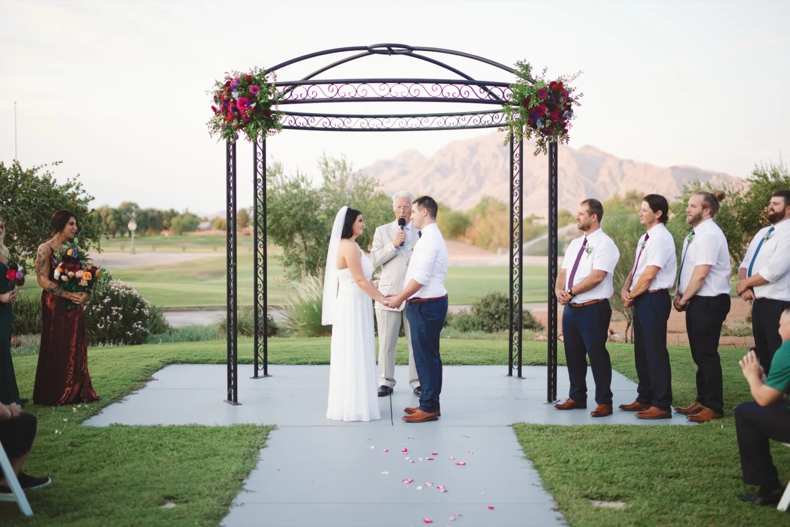 Couple holding hands under a black wrought iron rotunda on the ceremony lawn, the officiant between them, groomsmen lined up to one side and a bare mountain ridge across the golf course behind