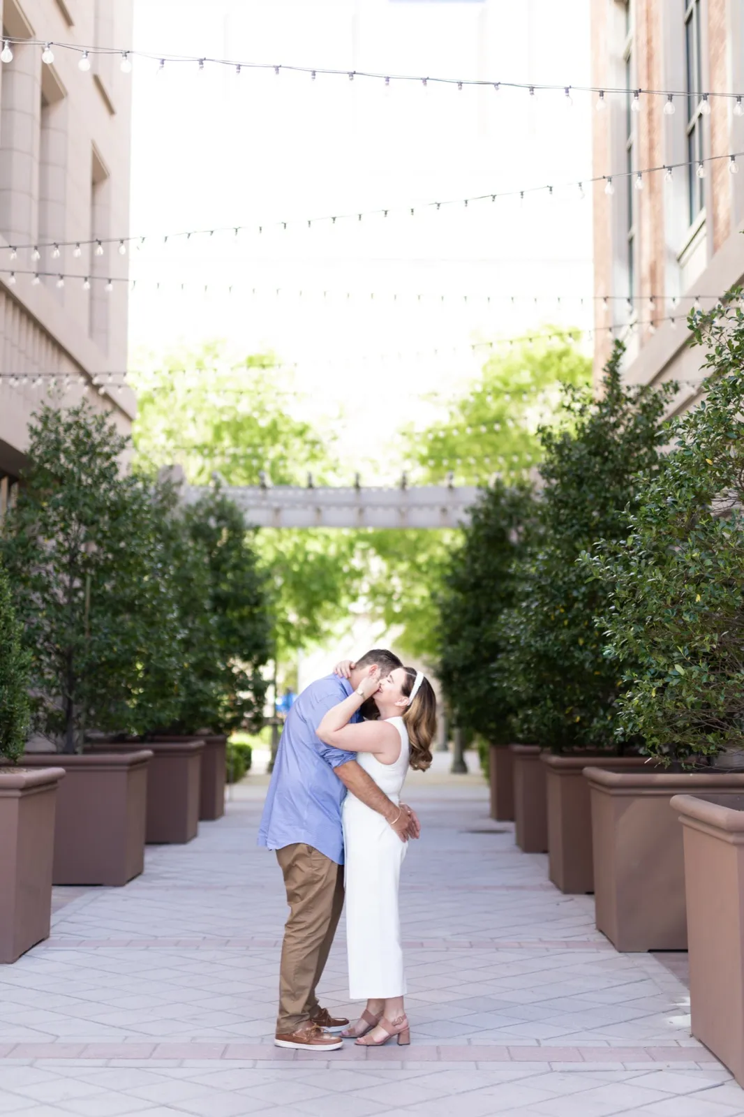 Couple in casual clothes kissing on the planter lined walkway under strings of cafe lights, green trees on both sides