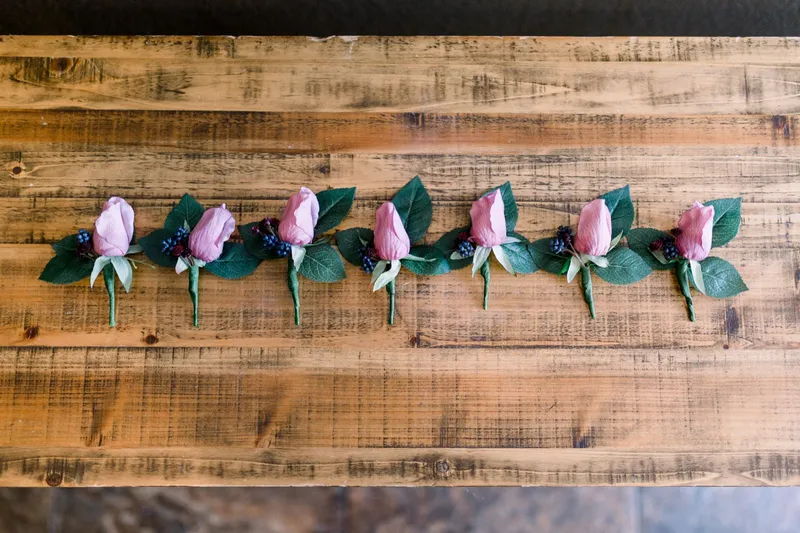 A row of pink rose boutonnieres laid out in a line on a rustic wood table before the ceremony