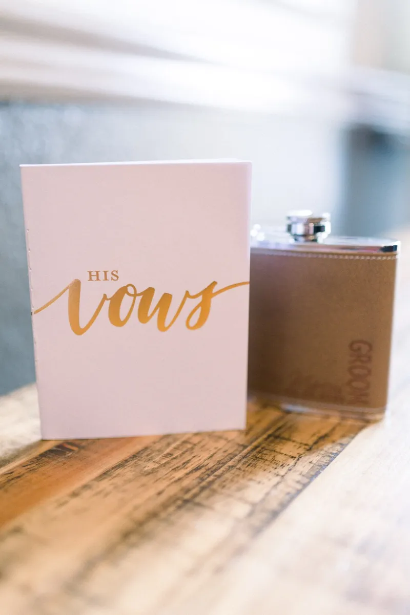 A blush book reading His Vows sitting next to a leather flask on a wood table in the getting ready room
