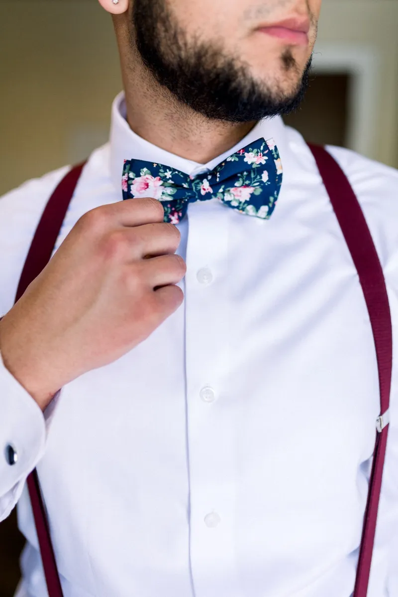 A groomsman in a white shirt and burgundy suspenders reaching up to straighten his navy floral bow tie