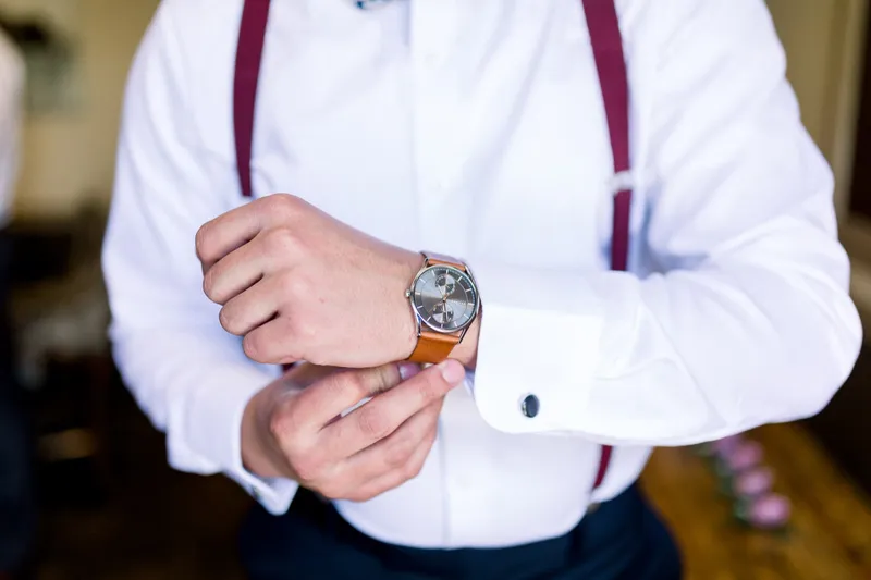 Close view of a groomsman fastening his watch, burgundy suspenders running over his white shirt