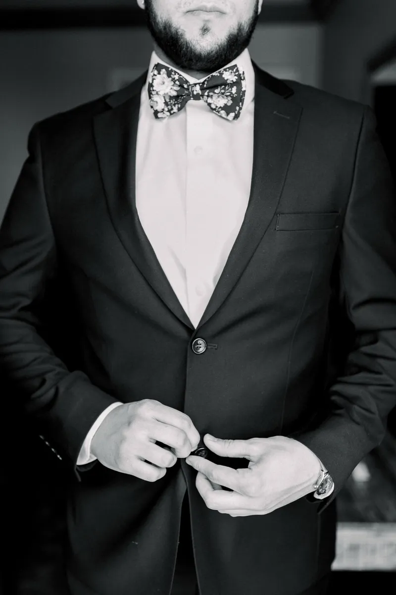 Black and white close view of the groom buttoning his suit jacket with his bow tie already tied