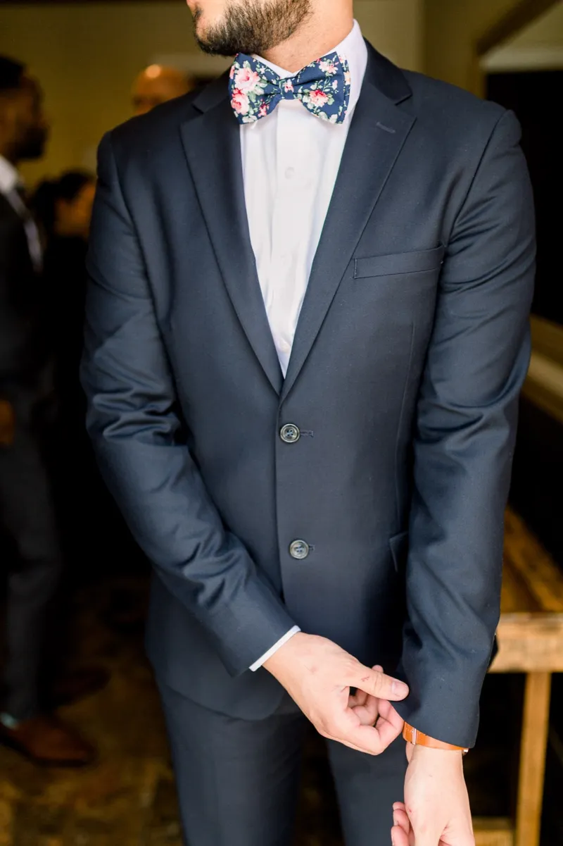 The groom in a navy suit and a navy floral bow tie pulling his shirt cuff straight before the ceremony