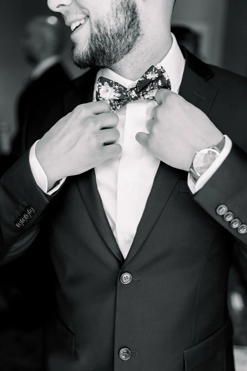 Black and white photo of the groom smiling with his head turned as he straightens his floral bow tie