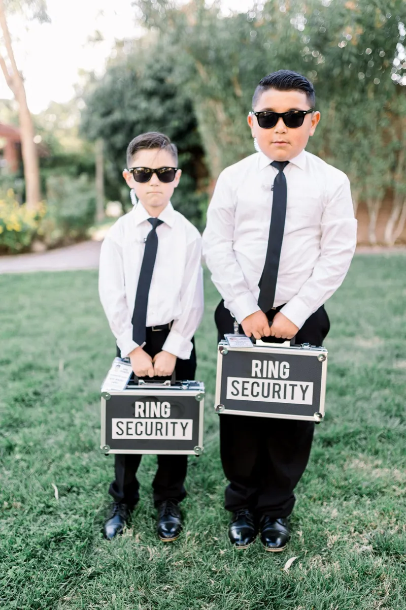 Two young ring bearers in sunglasses and black ties standing on the lawn holding metal cases labeled Ring Security