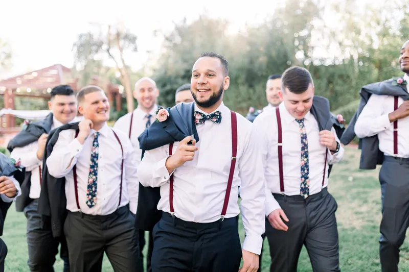 The groom laughing on the lawn with his groomsmen in white shirts and burgundy suspenders around him