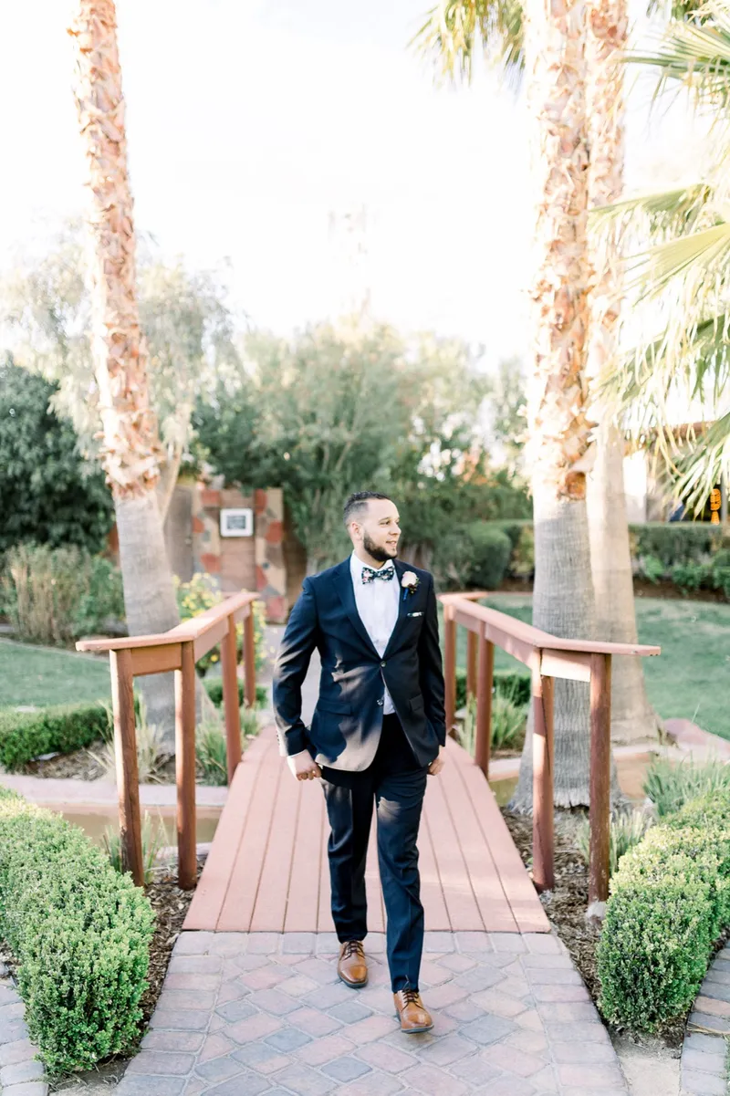 The groom in a navy suit walking toward the camera across the venue's wooden footbridge with palms on both sides