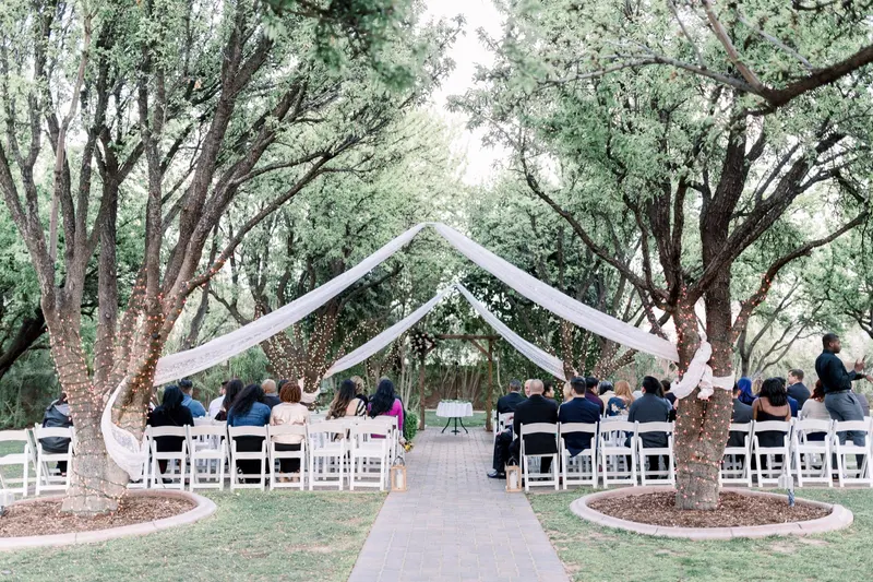 Guests seated on white chairs under lace drapes and string lights strung between the trees at an outdoor ceremony