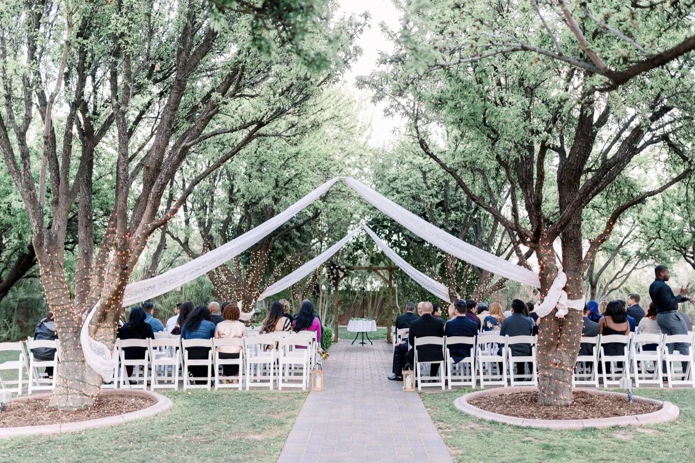 Guests seated on white chairs under lace drapes and string lights strung between the trees at an outdoor ceremony