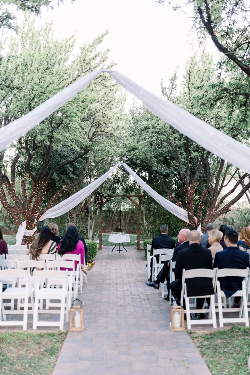 Looking up the paved aisle between rows of white chairs toward the draped trees and the wooden arbor