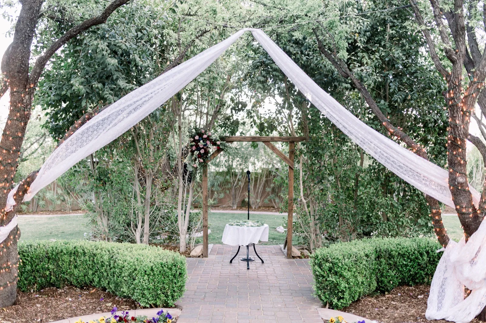 An empty garden ceremony site: lace drapes tented between trees, a rustic wooden arbor and a small unity table