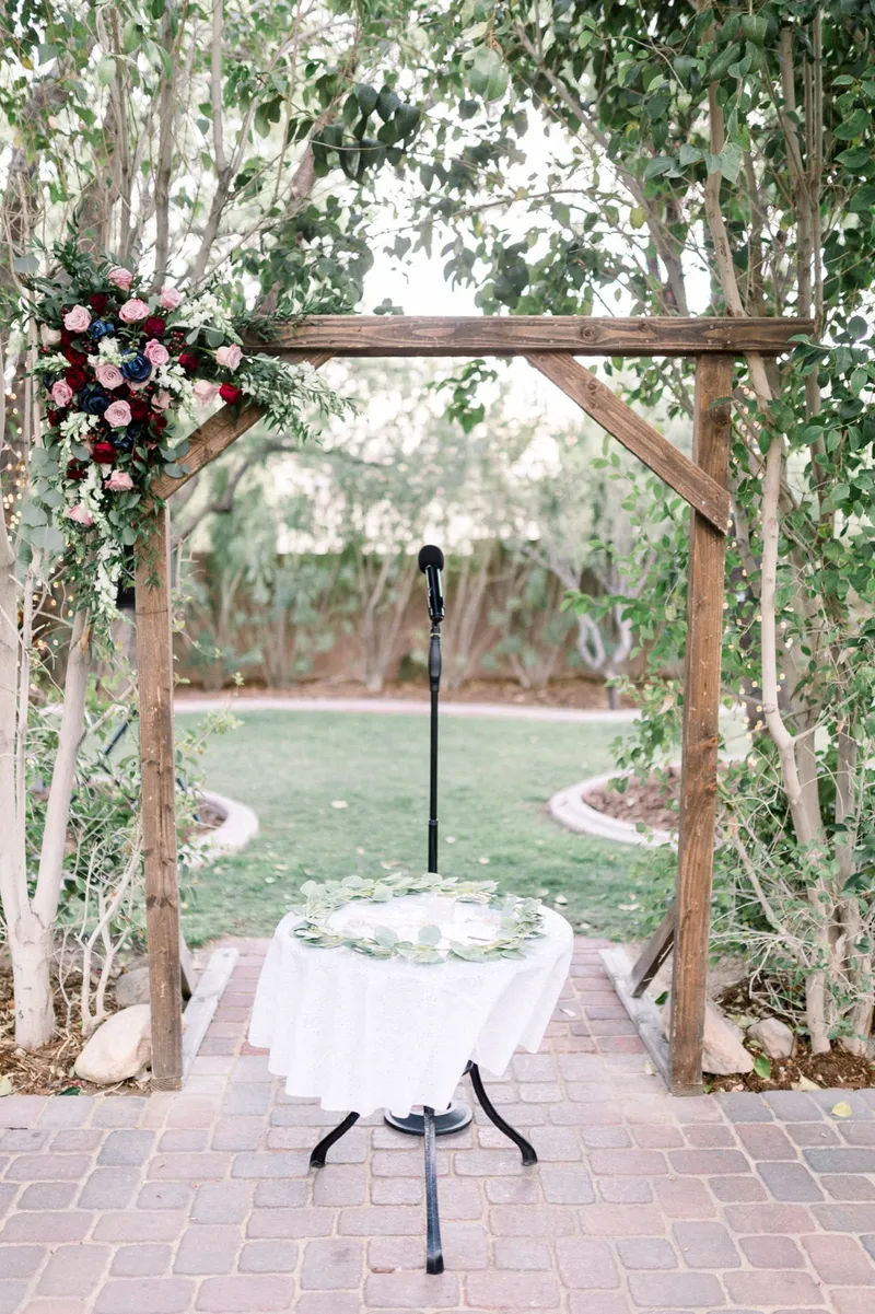 The wooden ceremony arbor with a burgundy and blush floral piece on one corner, a microphone stand and a small draped table underneath