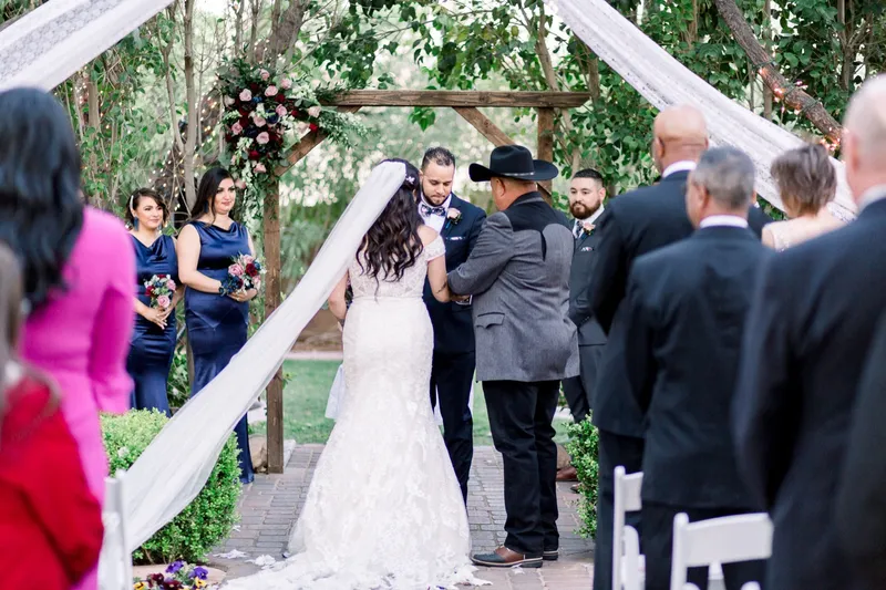 An older man in a cowboy hat handing the bride to the groom at the arbor, her long veil sweeping across the pavers behind her