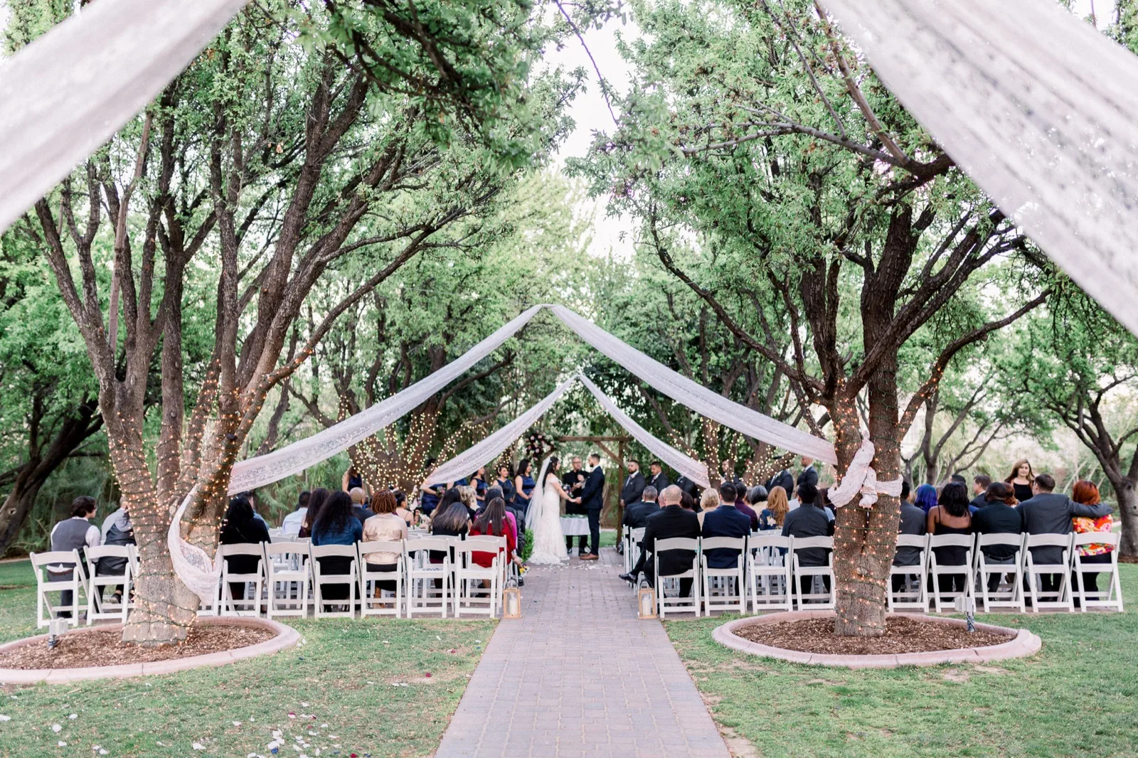 Wide view of an outdoor ceremony in a grove of trees, white folding chairs on both sides of a paver aisle and long lace panels strung up between the trunks over the guests