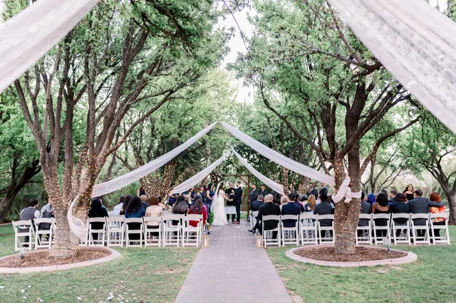 Wide view of an outdoor ceremony in a grove of trees, white folding chairs on both sides of a paver aisle and long lace panels strung up between the trunks over the guests