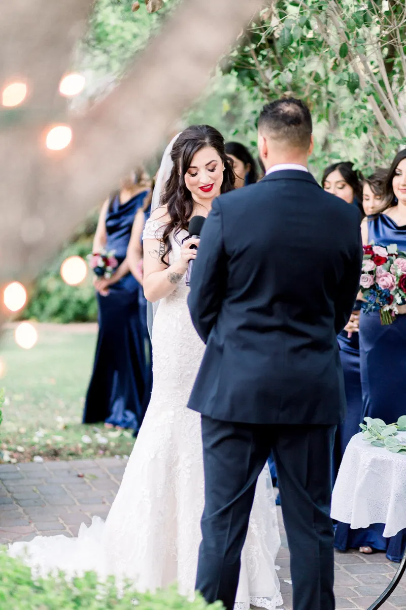 The bride reading her vows into a handheld microphone with the groom facing her and the string lights glowing behind