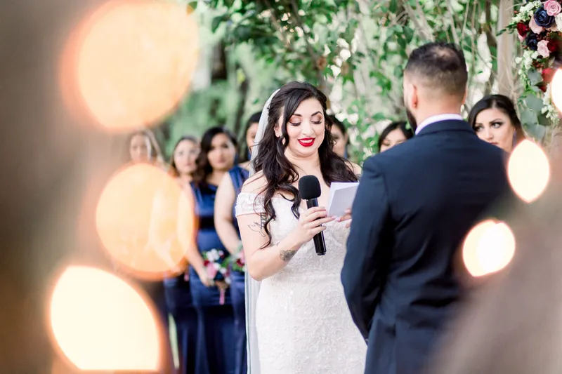 The bride speaking her vows with warm out of focus string lights scattered across the front of the frame