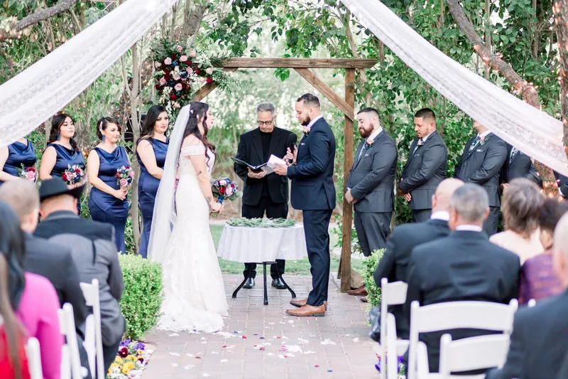 The officiant reading from a folder between the bride and groom at the arbor while guests watch from both sides