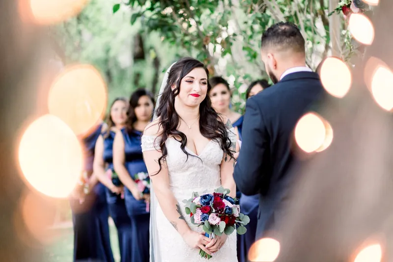 The bride holding her burgundy and navy bouquet during the ceremony, glowing lights blurred around her