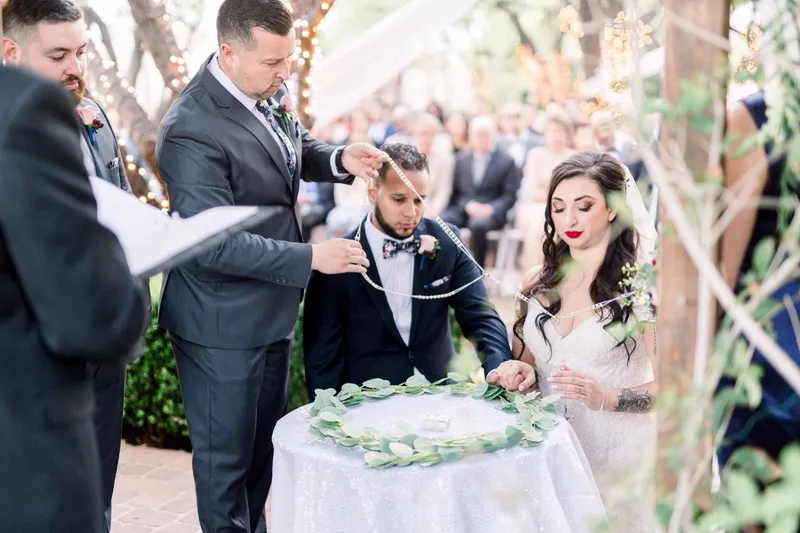 A beaded lazo cord being lowered over the couple's shoulders at a small table ringed with eucalyptus