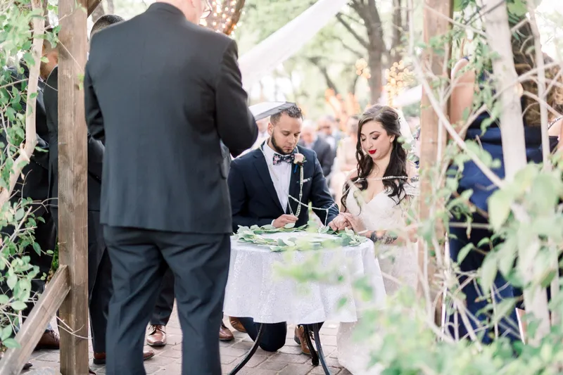 The officiant with his back to the camera as the couple stands together at the greenery ringed table
