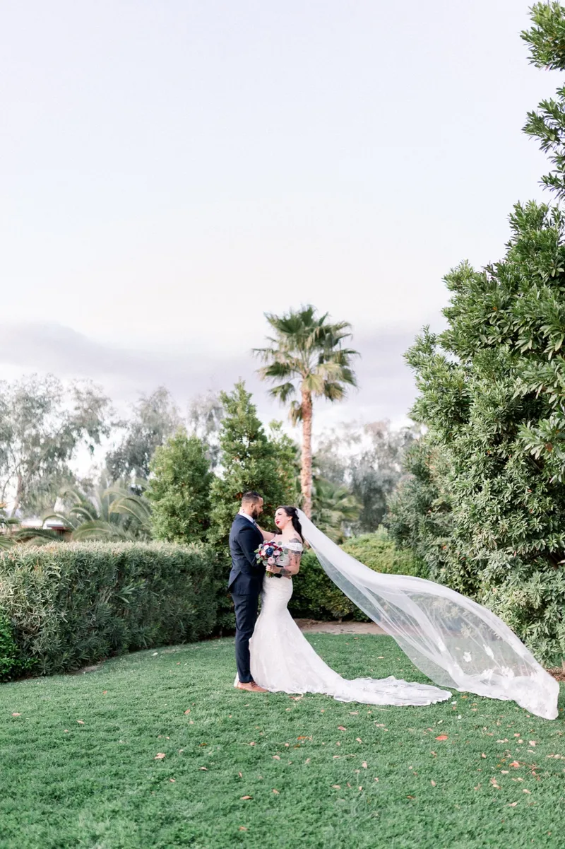 The couple on the venue lawn with the bride's long veil trailing across the grass, hedges and palms behind them
