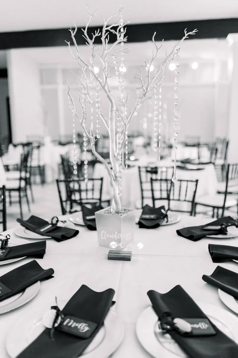 Black and white photo of a reception table set with dark napkins around a lit white branch centerpiece